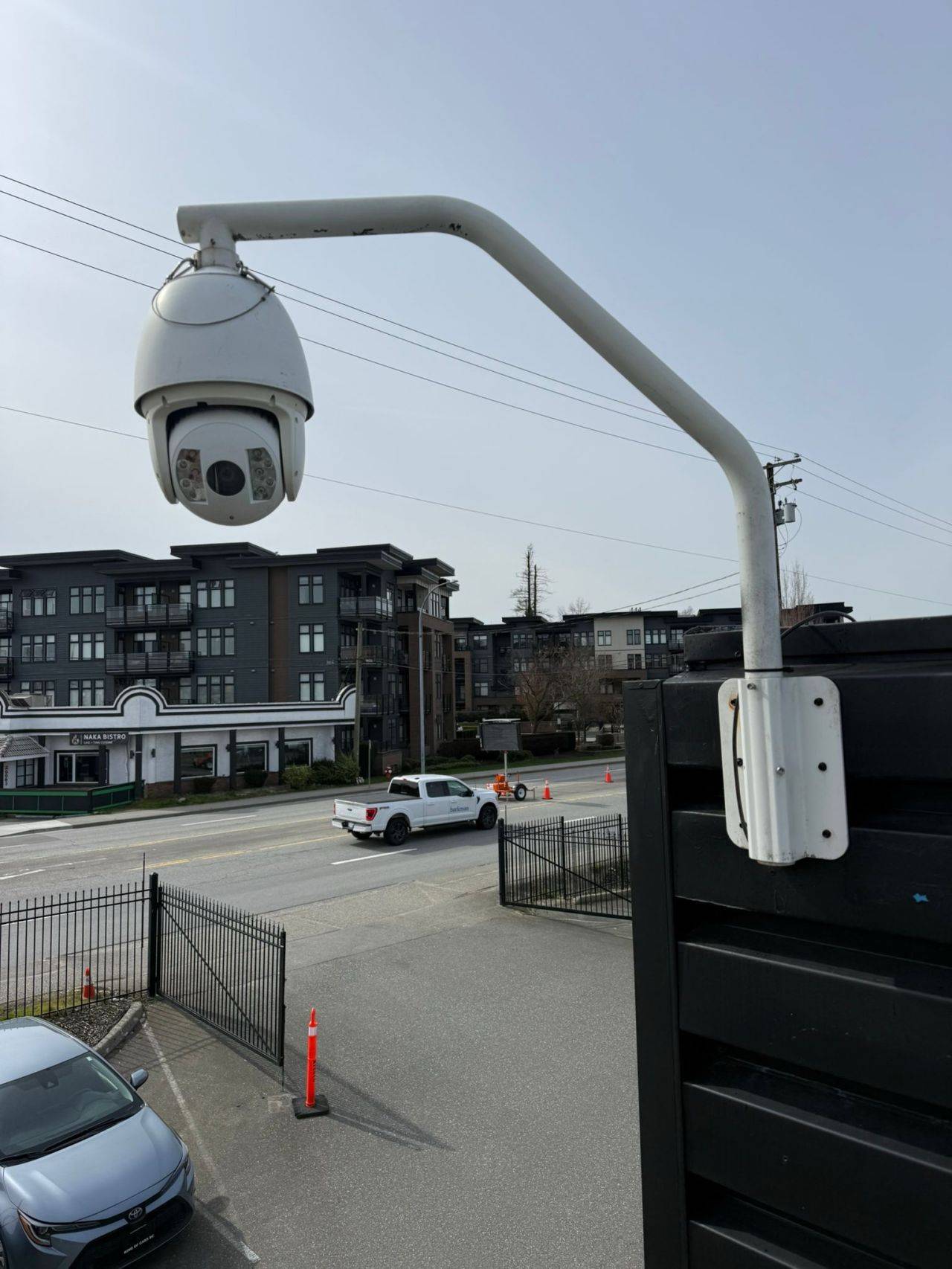 A dome camera on a curved metal arm above a paved entrance, with cars and buildings behind it